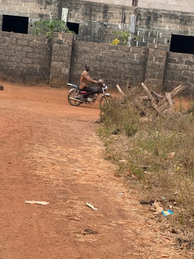 The image depicts an outdoor scene in Agbogugu, Nigeria, featuring a dark-skinned adult male on a motorcycle. The man, wearing a patterned long-sleeved shirt, black trousers, and dark shoes, is positioned on a silver-colored motorcycle, facing right. He appears to be in the process of operating or preparing to operate the vehicle on a reddish-brown dirt path. The path shows areas of dry, light-colored fibrous vegetation. To the immediate right, a dense patch of dry green and brown vegetation contains a pile of interlocking tree stumps or logs and scattered debris including plastic wrappers. In the background, a dark gray concrete block wall with several unglazed rectangular openings dominates the scene. Above this wall, an upper section of a building, also of concrete block construction, is visible. Green foliage, possibly from a papaya plant, grows near and over the wall. The environment suggests an unpaved, semi-rural or developing urban area.