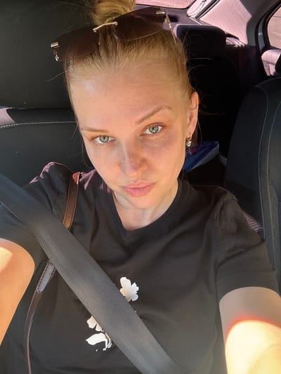 In the city of Fedorino, Russia, a young woman with blonde hair pulled into a messy bun, wearing sunglasses perched on her head, is captured in a selfie taken inside a car. She is wearing a black t-shirt with a white floral print and a brown strap from a bag is visible. A seatbelt is fastened across her chest. Her expression is neutral, and her green eyes are looking directly at the camera. The background shows car seats and a window with sunlight streaming in, suggesting it is daytime.