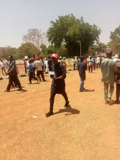 The image depicts an outdoor scene in Kagoma, Nigeria, under bright, clear daylight. The ground is unpaved, consisting of reddish-brown earth with scattered small gravel and some white debris. In the background, green trees are visible, along with a low, light-colored wall or fence. A street lamp stands behind this wall.
Approximately 20-30 individuals are present, predominantly males, dispersed across the scene. In the foreground, a male subject walks from left to right, looking forward. He wears a dark long-sleeved tunic, dark trousers, and dark shoes. A red baseball cap is on his head. He holds a small, white rectangular object in his left hand and a dark rectangular object, consistent with a smartphone, in his right hand. His shadow is cast distinctly to his right.
To the left and slightly behind him, another male walks right, wearing a patterned dark shirt, dark trousers with white stripes, and sandals. His shadow is also visible. To the right, another male subject is seen from the back, wearing a light green and white patterned garment with matching trousers.
Other individuals are scattered throughout the mid-ground and background, wearing various casual and traditional clothing. Some are standing, others are walking. No specific interactions are clearly discernible, but the individuals appear to be moving or gathered in an open area. Shadows are harsh and defined, indicating direct overhead sunlight.