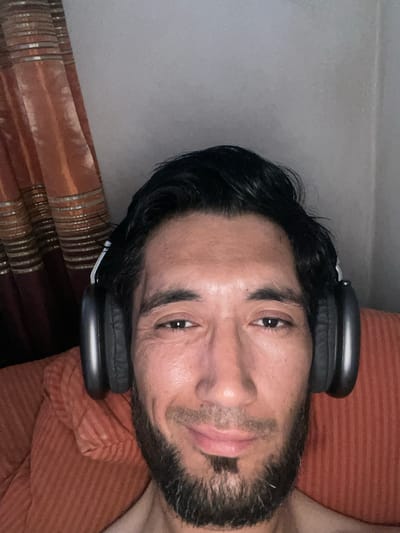 The image is a close-up, rotated selfie of a man wearing black headphones. He has dark hair, a beard, and is looking towards the camera with his eyes open. The man appears to be lying down on an orange, striped surface, possibly a couch or bed, with a brown, patterned fabric to his left. The lighting suggests it is indoors, and the time of day is unclear. There is no visible text in the image. The man's expression is neutral. The overall impression is casual and relaxed. The presence of headphones suggests he may be listening to music or engaged in some audio activity. The setting appears to be domestic.