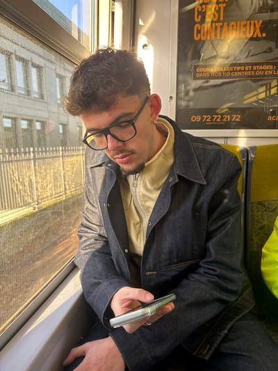 A young adult male is seated inside a train car, looking down at a smartphone held in his right hand. The individual has dark, curly hair, a light mustache, and a short beard, wearing black-framed glasses, a beige zippered high-collar garment, and a dark blue denim jacket. A silver ring is present on his right ring finger, and a small silver hoop earring is visible in his left ear.
The train interior features yellow-green patterned seats to the right of the subject and white overhead panels with embedded lighting. A black advertising poster with orange and white French text, including the phrase "C'EST CONTACIEUX" and a telephone number "09 72 72 21 72," is affixed to the wall behind the subject.
Through the window on the left, an urban environment in Bourg-la-Reine, France, is visible. This includes a multi-story building with numerous rectangular windows and a metallic fence or railing situated between the train and the building. Bright, natural sunlight streams through the window, illuminating the left side of the train car and the exterior scene. The window glass exhibits streaks and reflections. The smartphone, encased in a clear cover, displays an active screen.