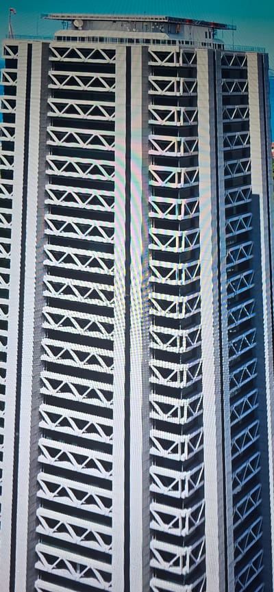 The image shows a close-up, aerial view of a modern, high-rise building, possibly under construction, with a distinctive facade featuring alternating dark and white geometric patterns. The building's exterior is characterized by repeating horizontal bands of dark panels interspersed with white structural elements that form triangular shapes. There are multiple vertical sections of the building visible, creating a strong sense of depth and scale. At the very top of the building, a flat roof structure is visible, equipped with antennas and other equipment, and a flag is flying from a pole. The building appears to be situated in an urban environment, as suggested by the blue sky and a hint of water or a body of land in the background on the right side of the image. There is no information about the city of Lippstadt, Germany, present in the image. No people or specific actions are discernible.
