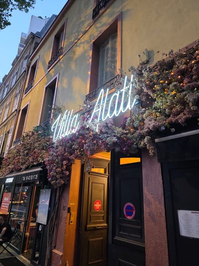 This is a medium shot, taken from a low angle looking up, of a building facade decorated with flowers and neon lights. The building appears to be a restaurant, as indicated by the "RESTAURANT" signage and a menu displayed on the window. The neon sign spells out "Alati" in bright white light. The flowers, in shades of pink and purple, are intertwined with small, warm-toned lights, suggesting an evening or nighttime setting. The building's facade is yellow, with dark brown doors and window frames. A round red and blue sign with text in French, indicating no parking or private property, is visible on one of the doors. Another sign displays a phone number: "Tel: 01 42 53 17 25". The overall impression is festive and inviting.