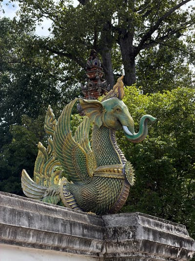 This media file depicts a close-up, low-angle shot of an ornate, mythical creature sculpture. The sculpture appears to be a fusion of an elephant and a bird-like creature with scales and wings, rendered in vibrant green and gold. The elephant's head, with its trunk raised and curved, is prominent, complemented by a tusk. The creature's body is covered in intricate scale patterns, and large, feathered wings are spread outwards. The sculpture is attached to a weathered, fluted stone pillar on the right side of the frame, suggesting it's part of an architectural element, possibly a temple or shrine.

In the background, a dense canopy of lush green trees dominates the upper portion of the image, with some branches extending into the frame. The lighting suggests it is daytime, likely with dappled sunlight filtering through the leaves. The overall atmosphere is one of cultural artistry and spiritual ambiance, consistent with a location like Ban Ko Klang, Thailand, known for its Buddhist temples and traditional crafts. There is no visible text in the image. The focus is entirely on the detailed craftsmanship of the sculpture and its natural surroundings.