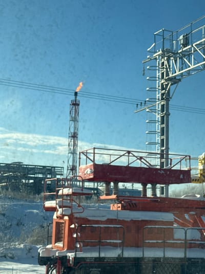 The image presents a forensic view of an industrial and railway environment located in Volkhov, Russia. A prominent, tall, red and white striped industrial flare stack is positioned in the left-center background, actively burning with an orange flame at its peak. To the left of the flare stack, an extensive industrial complex comprising multiple grey metal structures, platforms, and pipes is partially visible against a light blue sky. In the mid-ground to the right of the flare stack, a large grey metal railway gantry structure, featuring multiple support beams and electrical insulators, stands against the sky. Overhead electrical lines traverse horizontally across the upper portion of the frame, passing between the flare stack and the gantry. The foreground is dominated by a specialized orange and black railway vehicle, heavily coated with snow on its upper surfaces. This vehicle includes a distinctive elevated red work platform, also covered in snow, positioned above its main body. The ground throughout the scene is blanketed in white snow. The sky is predominantly clear blue with minimal cloud cover. No individuals are present in the scene.