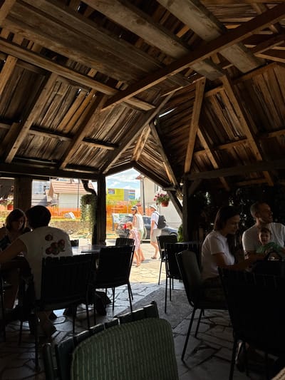 The image captures a lively outdoor seating area of a restaurant in Kolašin, Montenegro, on a sunny day. The open-air structure, constructed from rustic wooden beams and planks, provides shade and a natural ambiance. Several people are visible enjoying the setting: a woman and a man sit at a table on the right, the man holding a baby. In the center, a woman in a white shirt with a cherry design sits at a table, facing away from the camera. Another group of people, including a woman in a pink dress and a man in a white shirt and hat, walk in the background. The ground is paved with natural stone. Beyond the seating area, construction is visible with orange netting and buildings under construction, indicating ongoing development in the city. The scene suggests a casual and social atmosphere, likely a popular spot for dining and relaxation in Kolašin.