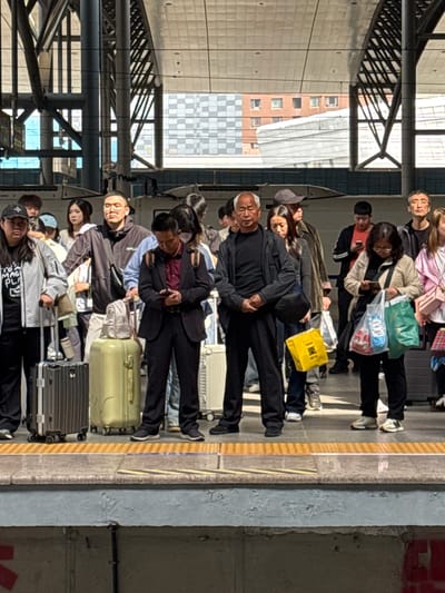 People stand in line on a train platform. The scene is brightly lit with sunlight streaming in from the building's frame. Several people have luggage. The location is likely Huanggu District, China.