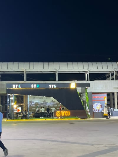 An exterior night-time, eye-level, medium shot captures a contemporary plaza area in Al-Ghizlaniya, Syria. The scene is dominated by a modern architectural structure featuring a pedestrian bridge overhead, supported by columns. Below the bridge, a dark, angled structure forms an entrance or facade. Illuminated signage is prominent; "STL," "STB" (with a blue-green hue), and "STA" are lit in white capital letters across a dark background. A large, bold yellow sign reads "STH" in capital letters, also illuminated.

Several individuals are present in the plaza. In the background, a group of people are gathered around tables and what appears to be a food or drink service area, with some seated. Further to the right, a few individuals stand near a display board featuring a globe graphic and Arabic text. One person in the foreground on the far left is partially visible, wearing dark pants and a light-colored shirt, walking away from the camera. The ground is paved with a light-colored, smooth material. Greenery is visible integrated into the dark, angled structure. The overall ambiance is one of an active, modern public space at night.