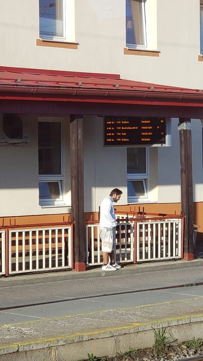 The image shows a man standing on a train platform in Spišská Nová Ves, Slovakia, looking down at his phone. He is wearing a white long-sleeved shirt and white shorts with a pattern on them, and white Adidas sneakers. Behind him, a digital display shows train departure times and destinations. The platform is covered by a red awning, and there is a white railing separating the platform from the tracks. The building behind the platform is light-colored with windows. The overall scene appears to be a sunny day at a train station.