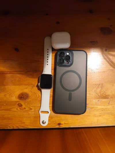 The image shows an Apple Watch, AirPods case, and an iPhone 14 Pro Max with a MagSafe case, arranged on a wooden surface. The Apple Watch has a white band and a gold-colored casing, with a black screen. The AirPods case is white and positioned above the iPhone. The iPhone is dark gray with a camera bump and a circular MagSafe indicator on the back. The wooden surface appears to be a desk or table, with visible grain and some minor imperfections. The lighting suggests an indoor setting, possibly a home or office. The photograph was taken in Neckertal, Switzerland.
