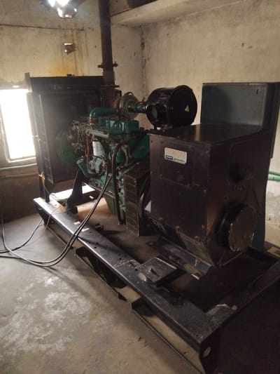 In an industrial room in Chhina Uttar, Pakistan, a large diesel generator is the primary subject. The generator, which appears to be a Stamford Power Generation unit, is dark green and black and sits on a black metal frame. It features a prominent black air filter on top, a large radiator on the left, and various hoses and electrical wiring connected to it. The room has concrete walls and floor. A window is visible on the left side of the image, letting in natural light. The ceiling has damage, with holes allowing light to enter. The overall scene suggests an operational or standby power source in a functional, albeit somewhat aged, industrial setting. There are no people visible in the image.