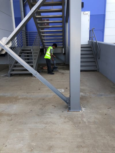 An outdoor scene in Hanak, Saudi Arabia, depicts a male individual sweeping a concrete ground area. The person, wearing a grey t-shirt, a high-visibility lime green safety vest, dark trousers, and dark shoes, is positioned slightly left of center, bent forward, actively sweeping with a dark dustpan and a long-handled brush or broom. A small pile of debris and a white bag are visible near his feet on the dirty concrete surface, which exhibits stains and striations. In the background, two sets of grey metal outdoor staircases ascend against a building featuring a blue facade. The left staircase ascends upwards and to the left, while the right staircase leads to a white emergency exit door on the blue and white wall. A dark rectangular sign with white Arabic text and an icon is mounted on the blue wall between the two staircases. Prominent grey metal structural elements include a vertical support beam near the center and a diagonal beam extending from the lower left, both bolted to the concrete ground.