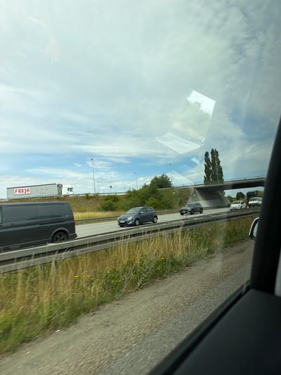 A highway scene is depicted from inside a vehicle. The focus is on the road ahead, with several vehicles visible. On the left, a dark grey van is in the foreground, moving on the highway. To its right, a dark grey sedan is also in motion. Further ahead, a white sedan and a light-colored van are visible. A large truck with a white trailer displaying the "FREJA" logo is in the distance on the left side of the highway. A bridge arches over the highway in the background, with tall trees visible behind it. The sky is partly cloudy with patches of blue. The surroundings include grassy areas and vegetation alongside the highway. The image appears to have been taken from a moving vehicle, suggesting a journey through the city of Karlslunde, Denmark.