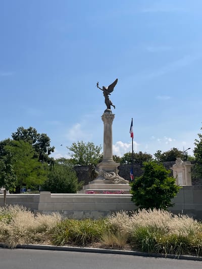 The image captures a monument in the city of Meaux, France. A bronze statue of a winged figure, likely representing victory or an angel, stands atop a tall, classical column. The figure holds a torch aloft. At the base of the monument, sculpted figures and reliefs are visible, along with a bed of pink flowers. Two French flags, one larger and one smaller, fly from a flagpole to the right of the main statue. The monument is situated in an outdoor setting with lush green trees and vegetation. In the foreground, ornamental grasses add texture and softness to the scene, with a grey paved area and a road in the lower section. The sky above is a clear blue with a few wispy clouds. The overall impression is one of a memorial site bathed in bright sunlight.