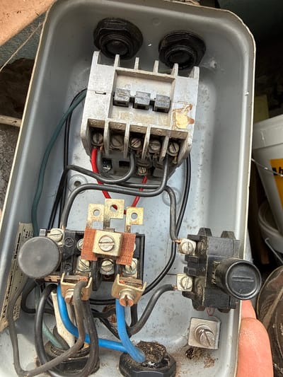 The image depicts the interior of an open, rectangular, light gray metal electrical box. Inside the box, there are multiple electrical components. On the left is a gray, multi-terminal block with multiple screws securing wires. Wires of various colors (black, red, green) connect to this block. Another black component with a cylindrical knob on top is located in the upper right corner of the box, with wires connected to it. In the lower right is a second terminal block, brown and black in color, with screws securing various wires including blue. There are black cord grips on the exterior sides of the box. The electrical components appear to be corroded. The image was taken near Sadievo, Bulgaria.