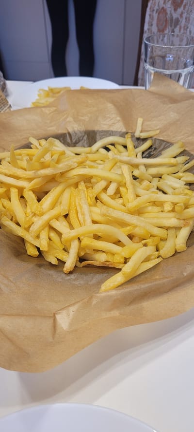 The image shows a basket of french fries on a table. The fries are golden brown and piled high in the basket, which is lined with brown paper. In the background, a glass of water can be seen, as well as part of a person's legs. The lighting suggests an indoor setting, likely a restaurant or dining area. There is no visible indication of the specific location within Dietzenbach, Germany.