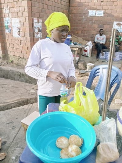 A woman wearing a yellow headscarf and white shirt stands outside on a sunny day, holding a water bottle. A blue tub filled with small, round baked goods is prominent in the foreground. A yellow plastic bag and other containers surround the tub. Behind the woman, a brick wall is adorned with posters. In the background, people are seated on chairs near a structure with a grocery bag. A blue plastic chair is also visible.