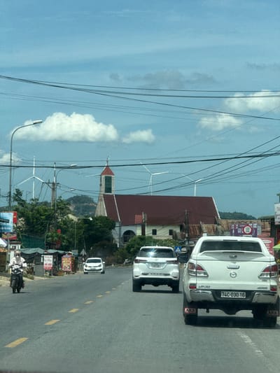 The image depicts a roadside scene in Tân Lập Commune, Vietnam, characterized by a bright, clear sky and scattered clouds, suggesting it is daytime. The road is paved and marked with yellow lines, indicating it's a thoroughfare. Several vehicles are visible, including two white pickup trucks and a white car. The first pickup truck, in the foreground, has a license plate reading "74C-030.16" and "ARB" decals on its mud flaps. Behind it, another white car displays a license plate "74A 161.00". A motorcycle with a rider is also present, partially obscured.
The surroundings are a mix of urban and natural elements. Buildings, some with red tiled roofs, line the road. Signs with Vietnamese text, such as "TÓC NAM - NỮ" (Men's - Women's Hair) and "OANH VÙ COM" (a food-related phrase), are visible, providing local context. In the background, lush green trees and a hillside can be seen. Notably, several wind turbines are present in the middle ground, suggesting a rural or semi-rural area with renewable energy infrastructure. Power lines crisscross the sky, a common sight in many regions. The overall impression is a bustling but somewhat rural roadside environment in Vietnam.