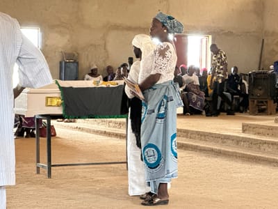 The image depicts a funeral service inside a building with rough, unplastered walls and a dirt floor, possibly located in Kufana, Nigeria. In the foreground, a coffin draped with a dark cloth and green fringe rests on a metal stand. To the right of the coffin, a woman wearing a blue headscarf and a white lace top with a patterned blue skirt is standing, holding a booklet. Behind her, other attendees, including men and women in traditional attire, are seated. To the far right, a large speaker is visible. A man in a patterned shirt is walking across the scene. The environment appears to be a community hall or church.