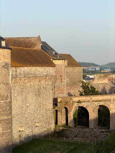 This image depicts a historical stone structure, possibly a fortress or castle, in Dieppe, France. The perspective is from an elevated vantage point, looking down and across the extensive stone buildings and their tiled roofs. The stone masonry is varied, with larger, rough-hewn blocks forming the main walls and smaller, more uniform stones used for detailing and pathways. Several arched openings are visible in one section of the building, suggesting an older architectural style.
The scene is bathed in natural light, indicating it was taken during the daytime, likely late afternoon or early morning given the warm, golden tones on the stone. The sky is clear and pale blue, suggesting fair weather. Beyond the immediate structures, a distant landscape unfolds, featuring rolling hills with scattered buildings and trees, hinting at the surrounding town or countryside. A patch of green, well-maintained grass is visible to the right, possibly a lawn or park area adjacent to the fortress.
No people are visible in the image. The focus is entirely on the architecture and the vastness of the historical complex. The overall impression is one of enduring history and solid construction, with a sense of peace and quietude due to the absence of human activity. The weathered appearance of the stone and tiles suggests the age of the structure.