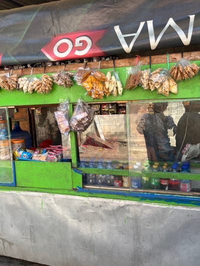 The image shows a street-side stall in Gedang, Indonesia, selling various snacks and drinks. The stall has a bright green wooden structure with a black tarp overhead displaying the white letters "GO MA". Several bags of snacks, including what appear to be fried banana chips and nuts, are hanging from the front of the stall. Inside the stall, shelves are stocked with bottled drinks, including water, soda, and other beverages. Some packaged snacks are also visible on a lower shelf. A large blue water dispenser is also present. The reflection in the glass of the stall shows a person, likely a vendor, wearing a dark patterned shirt, and some of the stall's interior. The stall is situated in an outdoor urban environment, with a metal paneling visible at the bottom of the image, suggesting it's part of a larger structure or building.