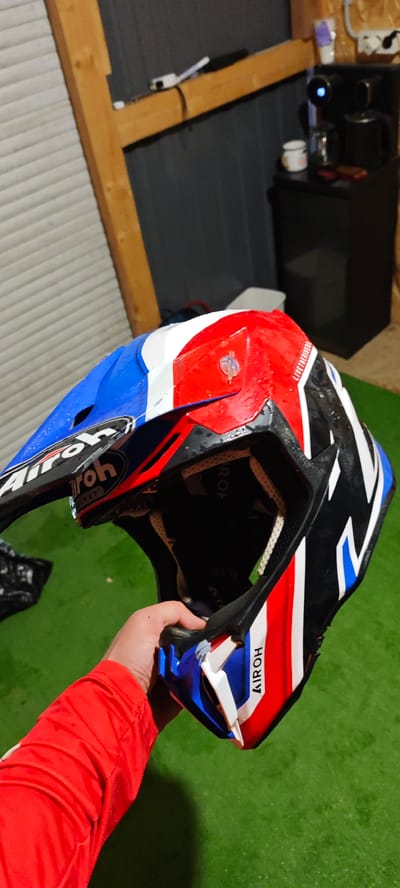 The image shows a person holding a blue, white, and red AIROH dirt bike helmet. The helmet has the AIROH logo prominently displayed. The person is wearing a red long-sleeved shirt. The background includes a green surface, possibly artificial turf, a wooden structure, and a black cabinet with a coffee maker on top. The location is Kazachiy Brod, Russia.