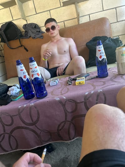 In Valea Mare, Moldova, a young man with short dark hair and sunglasses is seated on a brown leather couch, wearing black shorts and no shirt. He is holding a cigarette in his right hand and appears to be smoking it. On a table covered with a purple cloth, there are two blue bottles of "Blanc 1664" beer, a can of beer, a pack of Camel cigarettes, a mobile phone, and a pen. A black backpack is on the couch behind the man, along with a gray t-shirt. The scene is outdoors, with a brick wall in the background. The lighting suggests it is daytime.