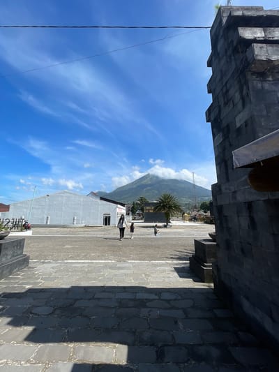 The image shows an outdoor scene in Kledung, Indonesia, with a view framed by the dark stone architecture of a building. In the distance, a mountain is visible, partially covered by clouds, set against a bright blue sky with wispy clouds. A building of light grey color, and with a simple design, is seen in the center, while two people are walking away from the camera across a paved area. A palm tree is also visible.