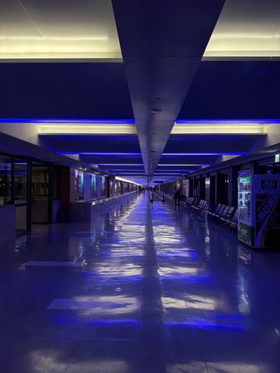 The image captures a long, indoor corridor in Taoyuan City, Taiwan. The corridor is illuminated with alternating white and blue vertical light fixtures along the left wall. The floor has a glossy surface, reflecting the blue light, creating a mirrored effect. Along the right side, there are rows of seats and vending machines. Several people are walking down the corridor in the distance.