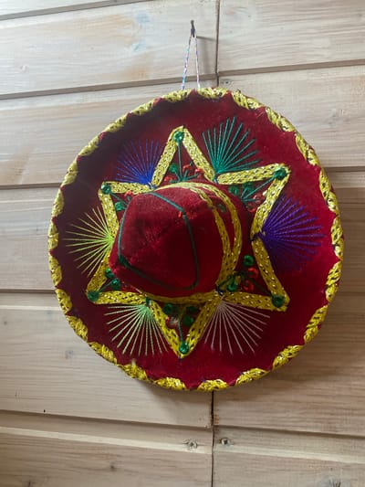 A red sombrero hat with yellow and green embroidery is hanging on a wooden wall. The hat is decorated with colorful threads in shades of blue, green, purple, and white, arranged in starburst patterns. Small green and red sequins are also present on the hat. The wall is made of light-colored wooden planks. The scene is indoors, likely in a residential or decorative setting. There are no people or animals visible in the image. The image is taken from a slightly elevated angle, looking down at the hat.
