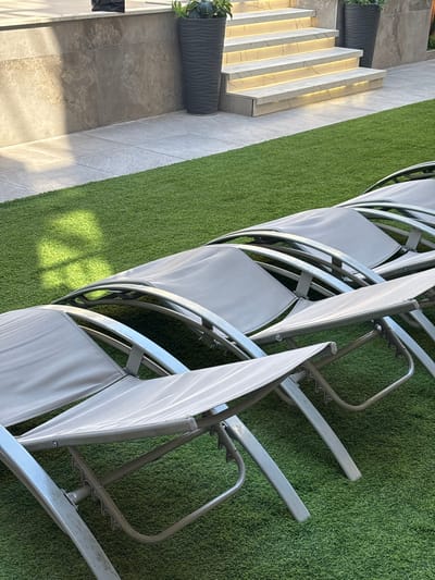 In Ayn al Basha, Jordan, an outdoor scene unfolds with artificial turf covering the ground. Several grey lounge chairs are neatly arranged in a row on the turf, their metal frames and fabric seats visible. Behind the chairs, a paved walkway leads to a set of illuminated outdoor stairs. The stairs are constructed with light-colored stone and have integrated lighting, casting a warm glow. To the left of the stairs, a wall made of large, grey stone slabs forms a backdrop, with a dark planter containing a green plant placed in front of it. To the right of the stairs, another dark planter with a similar plant stands. Sunlight casts a patterned shadow on the turf from an unseen source. The overall impression is of a well-maintained outdoor leisure area.