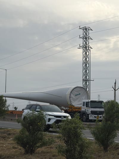 The image depicts a large, white wind turbine blade being transported on a flatbed truck. The truck, a white commercial vehicle, is driving on a paved road. A white SUV is also visible on the road, driving in the same direction as the truck. The scene is set against a cloudy sky, with a prominent electricity pylon in the background. Vegetation, including bushes and dry grass, lines the side of the road. The overall environment suggests an outdoor setting, possibly on the outskirts of a city or in a rural area. The presence of the wind turbine blade indicates a focus on renewable energy infrastructure. The location is identified as Sugalimetta, India. There are no people clearly visible in the image.