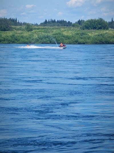 Two individuals are riding a jet ski on a broad, calm river. The person in front is steering the jet ski, while the person behind them is sitting. Both individuals are wearing bright orange life vests. The jet ski is creating a white wake as it moves through the blue water. In the background, a dense line of green trees and shrubs forms the riverbank, with a few taller trees interspersed. The sky above is partly cloudy with a few white clouds scattered across a pale blue expanse. The scene appears to be set in a natural, rural environment, possibly near Abalakovo, Russia, given the mention of the location.
