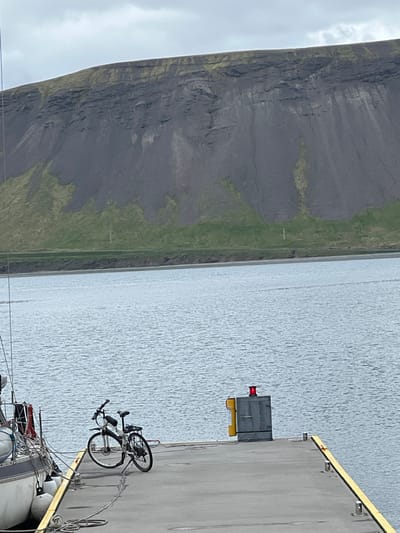The scene is a dock in Grundarfjörður, Iceland. A black bicycle is parked on the gray concrete dock with a yellow stripe along the edge. There is a gray metal box with a red light on top of it, located on the dock near the bicycle. In the background, a large gray cliff with green vegetation on top of it is visible. The water in front of the dock is calm and blue. The sky is overcast.