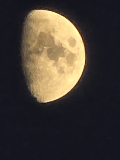The media file is a close-up, dimly lit photograph of the moon against a dark night sky. The moon is partially illuminated, showing a textured surface with visible craters and lunar features. Its color is a pale yellow-orange, suggesting it may be close to the horizon or affected by atmospheric conditions. The overall impression is one of a celestial body in space. There are no people or animals visible in the image, nor any text. The scene is a nighttime view of the moon. Given the location context of Montrouge, France, this is likely a view of the moon as seen from that area. The image appears to be taken at night, with no specific weather conditions discernible beyond the darkness of the sky.