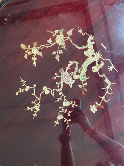 The image displays a close-up view of a dark, wooden surface adorned with intricate mother-of-pearl inlay. The inlay depicts a floral motif, featuring stylized branches, leaves, and flowers in a creamy, iridescent material. Some of the flowers appear to be in bloom, while others are represented as buds. Small, reddish accents are visible on some of the petals and leaves, adding subtle color variation. The wood itself has a rich, dark red-brown hue and appears to have a polished, glossy finish, showing numerous light scratches and some smudges, suggesting age and use.

Reflected on the surface, the arms and upper torso of at least two individuals are visible. One person's arms are bent and appear to be resting on or near the surface, with a bracelet visible on one wrist. Another reflection shows what could be part of a face and shoulder, also leaning into the frame. The reflections are slightly distorted, indicating the curved or uneven nature of the surface or the angle of the reflections. The lighting suggests an indoor environment, possibly with artificial light, given the reflections. The context of Quezon City, Philippines, doesn't provide specific visual cues within the image itself, but places the object within a particular geographical and cultural setting. No text is visible in the image.