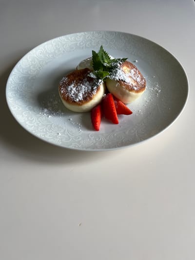 A close-up, overhead shot captures two syrniki, which are cheese pancakes, on a white plate with a raised, ornate floral pattern. The syrniki are golden brown and dusted with powdered sugar. Three slices of fresh strawberry are arranged to the left of the syrniki, and a sprig of mint garnishes the top. A dollop of cream is visible behind the syrniki. The plate rests on a light-colored, smooth surface, likely a table. The background is a plain, pale gray wall. The image is lit by natural light, creating soft shadows. This scene is located in Otyakovo, Russia.