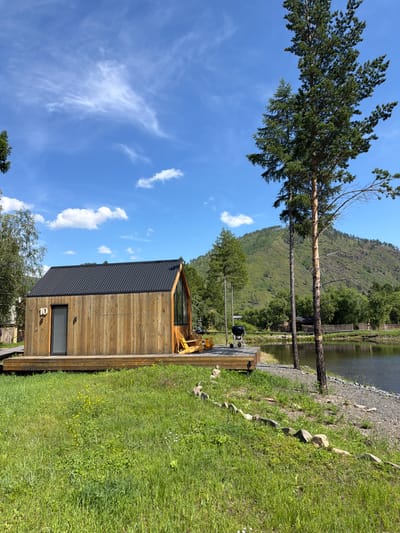 The image depicts a wooden cabin with a black metal roof, situated on a wooden deck next to a body of water. The cabin has a large glass window on its right side and a door on its left side, with the number "10" above the door. Two yellow chairs are placed on the deck in front of the glass window. A black grill is also visible on the deck. The scene is set in a natural environment with lush green grass, trees, and a large hill in the background. The sky is blue with scattered white clouds. The location is identified as Bol'shoe Goloustnoe, Russia.