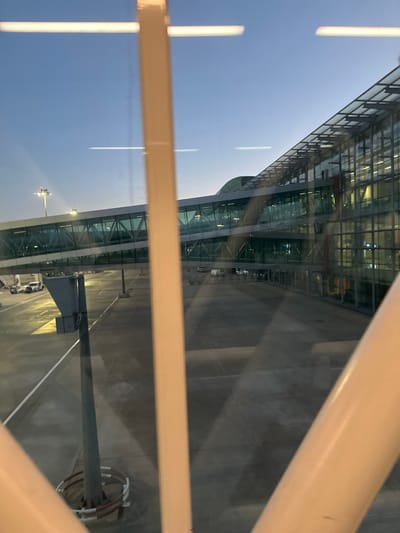 The image depicts an outdoor view of Adnan Menderes Airport (ADB) in Gaziemir, Turkey, observed from inside a building or jet bridge through a window, partially obscured by light-colored structural beams in the foreground. The sky above is clear, transitioning from light blue at the top to a lighter hue near the horizon, indicating twilight conditions. Artificial lighting is active throughout the scene.

On the right, a modern airport terminal building with a multi-story glass facade and a sloped glass roof is visible, with internal lights illuminating its interior. Extending from the terminal towards the left is an enclosed, elevated glass jet bridge, also illuminated internally. Below the jet bridge and in front of the terminal is a paved tarmac area.

On the tarmac to the left, a tall light pole with multiple luminaires provides illumination, and a white ground support vehicle, possibly an aircraft tug, is partially visible. Further into the lower-left, a grey, angular structure with a circular base marked with red and white stripes stands on the tarmac. Faint reflections of internal light sources appear as horizontal streaks on the viewing window. No distinct individuals are discernible, and no specific actions or interactions are depicted.