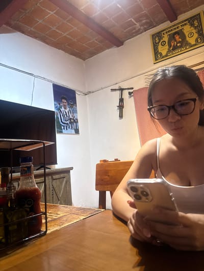 A young woman with glasses is holding a smartphone in her hands. She's seated at a wooden table. A rack with condiments sits on the table. Behind her, the room has brick and painted walls, posters, and other objects. This setting suggests an indoor location, possibly a restaurant or home in Chipilo de Francisco Javier Mina, Mexico.