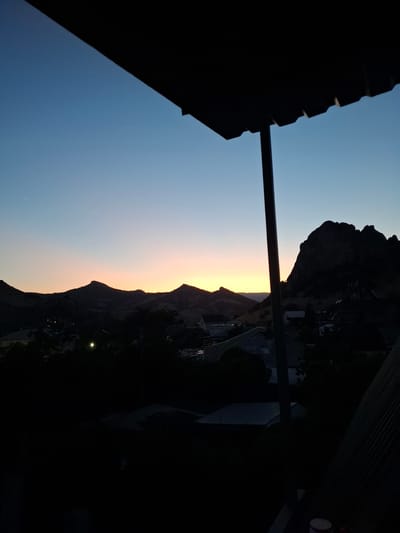 The image depicts a twilight scene from a balcony or covered area, looking out over a silhouetted landscape. The sky transitions from a deep blue at the top to a soft orange and yellow glow near the horizon, indicating the presence of a setting or rising sun. In the foreground, the dark silhouette of a modern roof structure with visible supports juts into the frame from the top and right. Below this, a dark, shadowed foreground obscures details of what appears to be a residential area with scattered buildings and trees. In the mid-ground, a range of hills or mountains is visible in silhouette against the lighter sky. One prominent, craggy peak stands out on the right side of the image. A few faint lights are visible in the mid-ground, suggesting the presence of human habitation. The overall impression is one of a peaceful, dusk or dawn view from a vantage point. The location is not specified as Щебетовский поселковый совет, Ukraine.