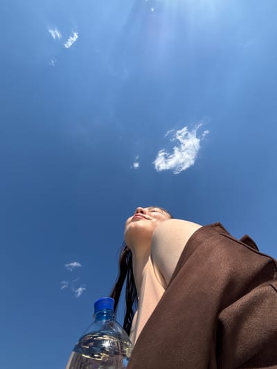 The image shows a low-angle view of a person holding a clear plastic water bottle against a bright blue sky with scattered white clouds. The person is wearing a brown garment. The top of the person's head and neck are visible. The scene is outdoors during daylight. There is no indication of the location being Zaosinovo, Russia.