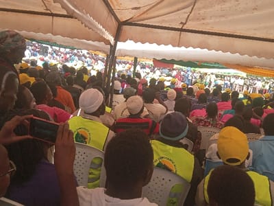 A large crowd gathers under a tent, many wearing yellow hats and vests. "UNITED DEMOCRATIC ALLIANCE" is printed on the vests. Attendees are seated in rows of white plastic chairs. A person in the foreground holds up a smartphone, presumably taking a photo or video of the event. A green, red, and yellow banner can be seen in the background.