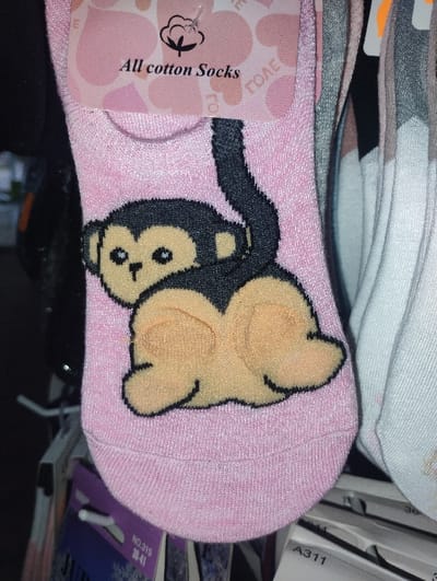 This image depicts a pink ankle-length sock, suspended from a retail display hook, within a commercial establishment in Byal Izvor, Bulgaria. The sock features a black and beige cartoon monkey design on its posterior, with the monkey positioned to show its rear towards the viewer, its head turned slightly left, and a black tail extending upwards. A pink cardboard tag, secured by a white plastic hanger, is attached to the sock's cuff, displaying the text "All cotton Socks" along with a cotton boll icon and partially visible Cyrillic text "ГОЛЕ". To the right, additional socks of varying colors (grey, white, multi-patterned) are visible, similarly displayed. Below the pink sock, portions of other product tags are discernable, one purple with "NO.315" and "36-41", and another white tag marked "A311". No people are visible; the scene is a close-up of merchandise, indicating a retail environment.