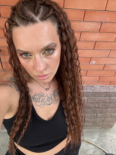 A woman with long, curly brown hair styled in braids stands in front of a brick wall. She is wearing a black tank top and has tattoos on her chest and arm. Her makeup includes bold eyeliner and green eyeshadow. She is also wearing a nose ring and a necklace with a small ball pendant. The brick wall appears to be an exterior wall, possibly in a backyard or courtyard setting. The lighting suggests it is daytime, and the overall scene is casual and informal. There is no visible indication of the specific location within Grushevskaya, Russia, beyond the presence of a brick wall, which is a common architectural element.