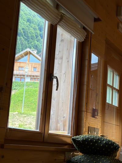 This is an interior shot looking out a window of a wooden cabin in Sauris/Zahre, Italy. The window frames are made of wood, and the walls are also paneled with wood. Outside, a green, tree-covered mountainside is visible, with a modern wooden house and a streetlamp in the foreground. The window is partially open, revealing the interior of the cabin, which includes a decorative pillow with green polka dots on a surface. To the right, another smaller window reflects the interior of the room and the wooden walls. A blind is partially rolled up above the main window.