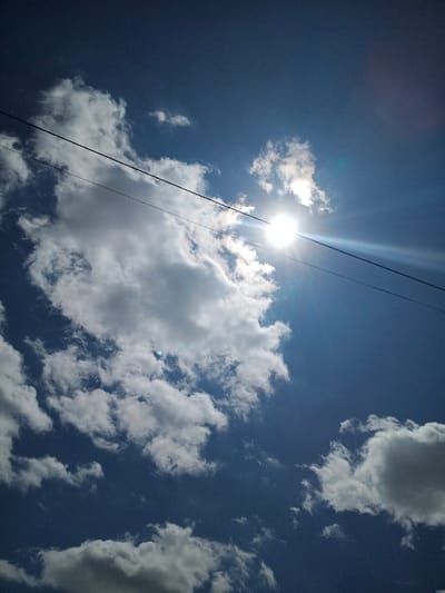The scene captures an upward perspective of the sky in Chitungwiza, Zimbabwe, under bright daylight conditions. The sky is predominantly deep blue, populated by scattered cumulus clouds ranging in color from white to light grey. The sun, a brilliant white orb, is positioned in the upper right quadrant, partially veiled by a cloud and generating a strong lens flare. Two dark, parallel utility lines are visible diagonally spanning the frame from the upper left to the lower right, crossing in front of the sun and clouds. No individuals, ground-level structures, or actions are depicted.