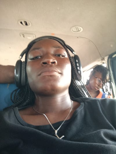 The image presents an interior scene within a vehicle, identifiable as a bus or commercial van, located in the city of Ibafo, Nigeria. The foreground is dominated by a young Black female, photographed from a low angle, wearing large black over-ear headphones. She has braided hair, a dark complexion, and is dressed in a black crew-neck t-shirt with a silver-colored chain necklace featuring an 'L' shaped pendant. Her gaze is directed towards the camera, with a neutral facial expression. Behind her, positioned to the right, another young Black female is partially visible. This individual wears glasses, appears to have braided hair, and is dressed in a garment with orange and blue coloration. The vehicle's interior features a light-colored ceiling with two overhead air vents and a light fixture. A blue fabric upholstery is visible on a seat to the left of the foreground subject, and a dark-colored curtain is partially visible on the right side of the frame, adjacent to the background subject.