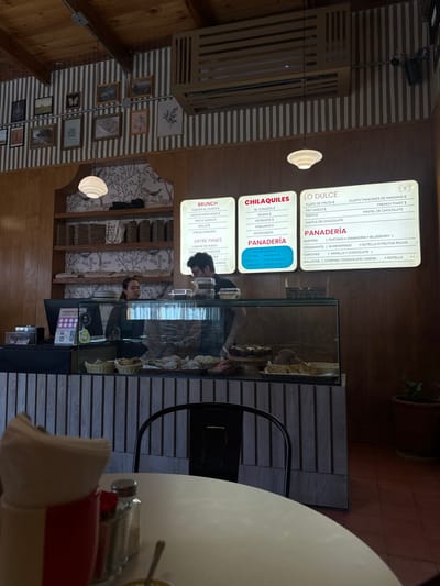 A restaurant menu board advertises "Brunch," "Chilaquiles," "Panaderia," and "Lo Dulce" options. Brunch includes eggs, Mac & Cheese, and sandwiches. Chilaquiles offers different sauces. The bakery selection features rolls, scones, brownies, and chocolate. Lo Dulce includes fruit plates, waffle, and muffins. "Panaderia" lists croissants, conchas, and cookies. The menu is illuminated and positioned above a pastry display counter, with two staff members seen behind it. A table with a chair and condiments occupies the foreground.