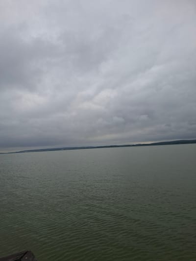 The media file depicts a wide, serene landscape dominated by a lake and a cloudy sky. The water, a murky green, occupies the right side of the frame and shows gentle ripples across its surface. A distant, dark shoreline is visible, separating the water from the sky. The sky, on the left side of the image, is filled with heavy, grey clouds, suggesting an overcast day, possibly late afternoon or early evening due to the subdued light. There are no visible people or specific objects in the scene, creating a sense of natural stillness. The location is identified as Зэльвенскі сельскі Савет, Belarus, implying a rural or natural setting. The overall mood is calm and perhaps a little somber due to the cloudy weather. No text is visible in the media file.